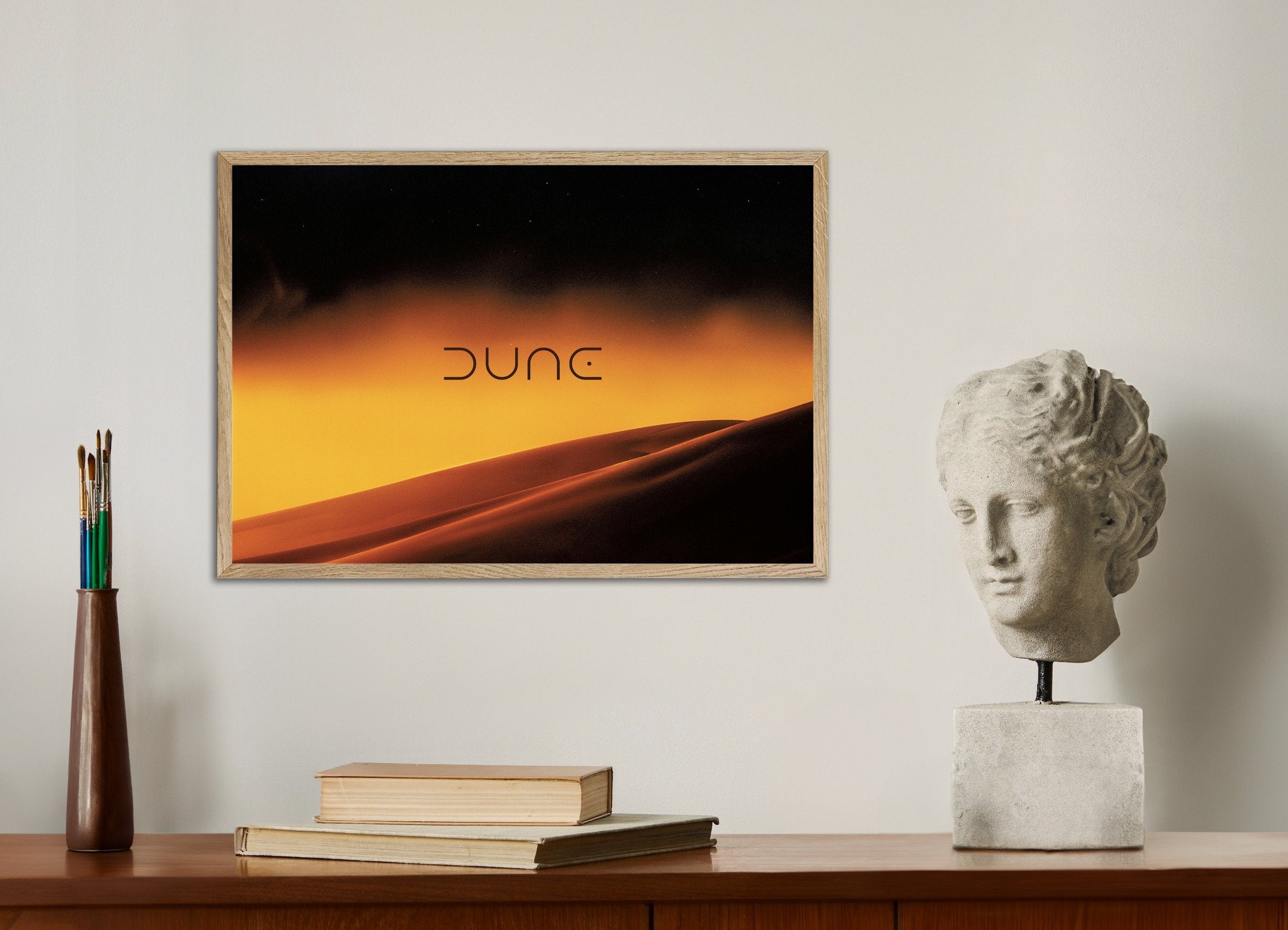 Framed artwork with 'Dune' title on a wall above a wooden desk with books and a sculpture.