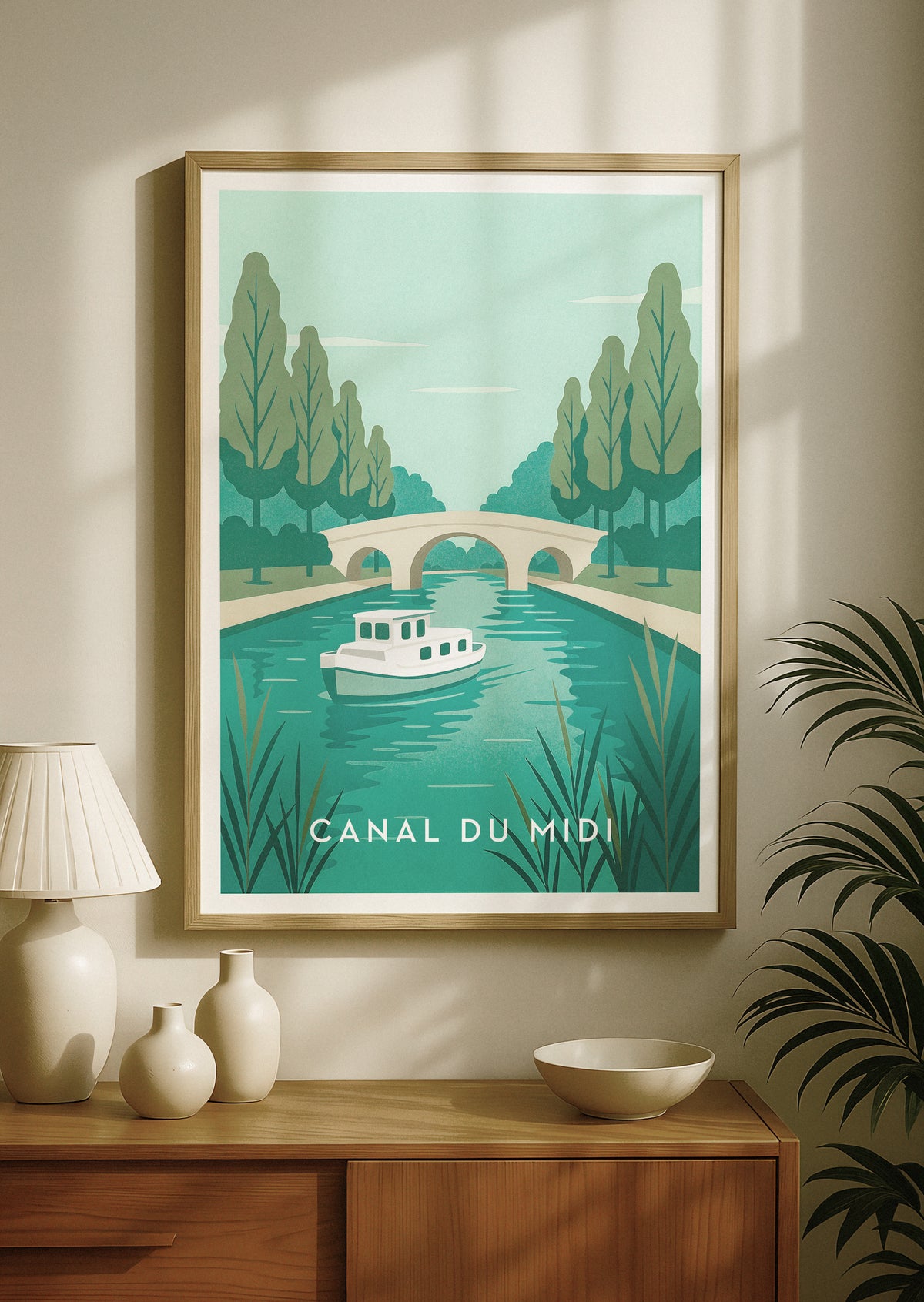 Ingelijste reisposter van het Canal du Midi in Frankrijk, met een retroboot op tealblauw water, bomen en een stenen brug. De poster hangt aan een beige muur boven een houten consoletafel met decoratieve vazen en een lamp.